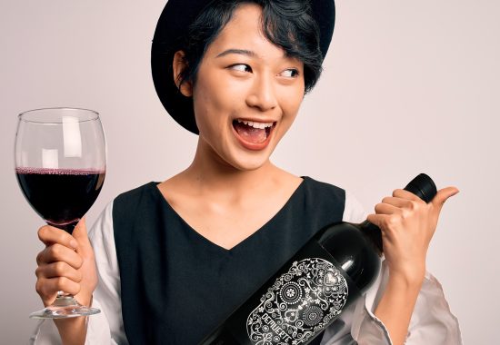 Young beautiful asian sommelier girl drinking glass of red wine over isolated white background pointing and showing with thumb up to the side with happy face smiling